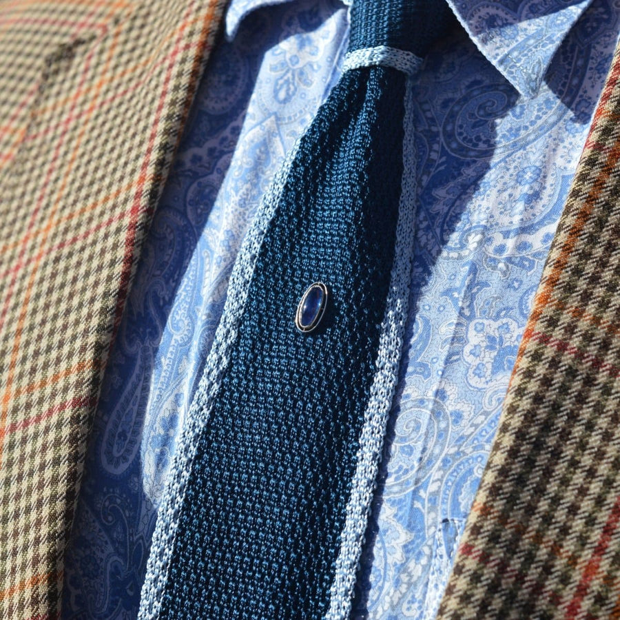 Art Deco platinum and sapphire tie pin on a textured navy tie, set against a patterned shirt and plaid blazer.