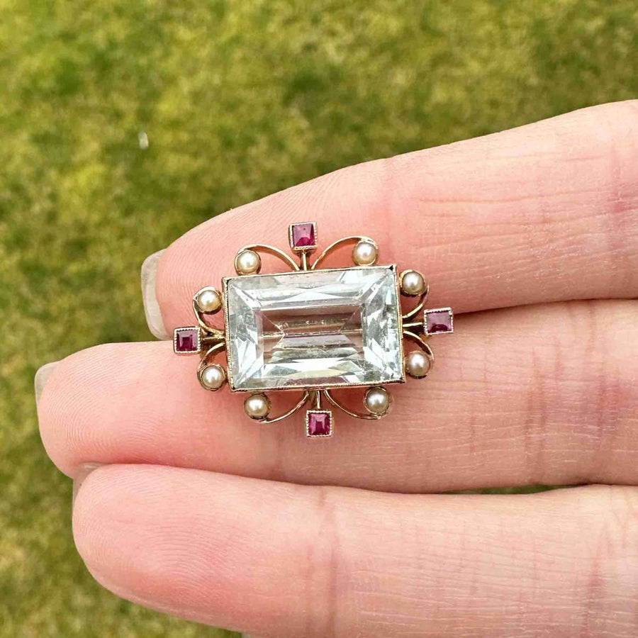 Edwardian 18ct gold brooch featuring aquamarine, rubies, and pearls, held in a hand with greenery background.