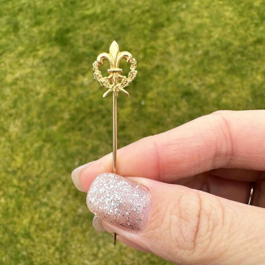 Edwardian 18ct gold French fleur-de-lis wreath tie pin held in hand against green background.
