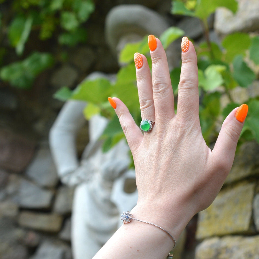 Hand with vibrant orange nails showcasing an Art Deco jadeite and diamond cluster ring in a garden setting.