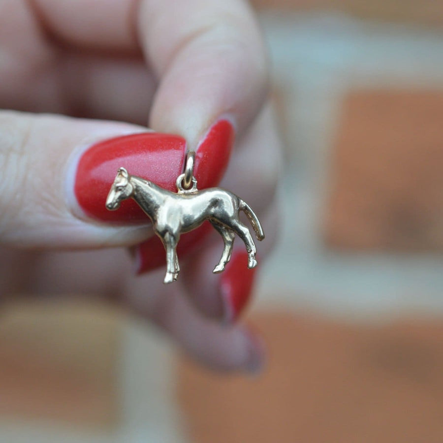 Vintage 9ct gold horse charm pendant held by a hand with red nails against a blurred background.