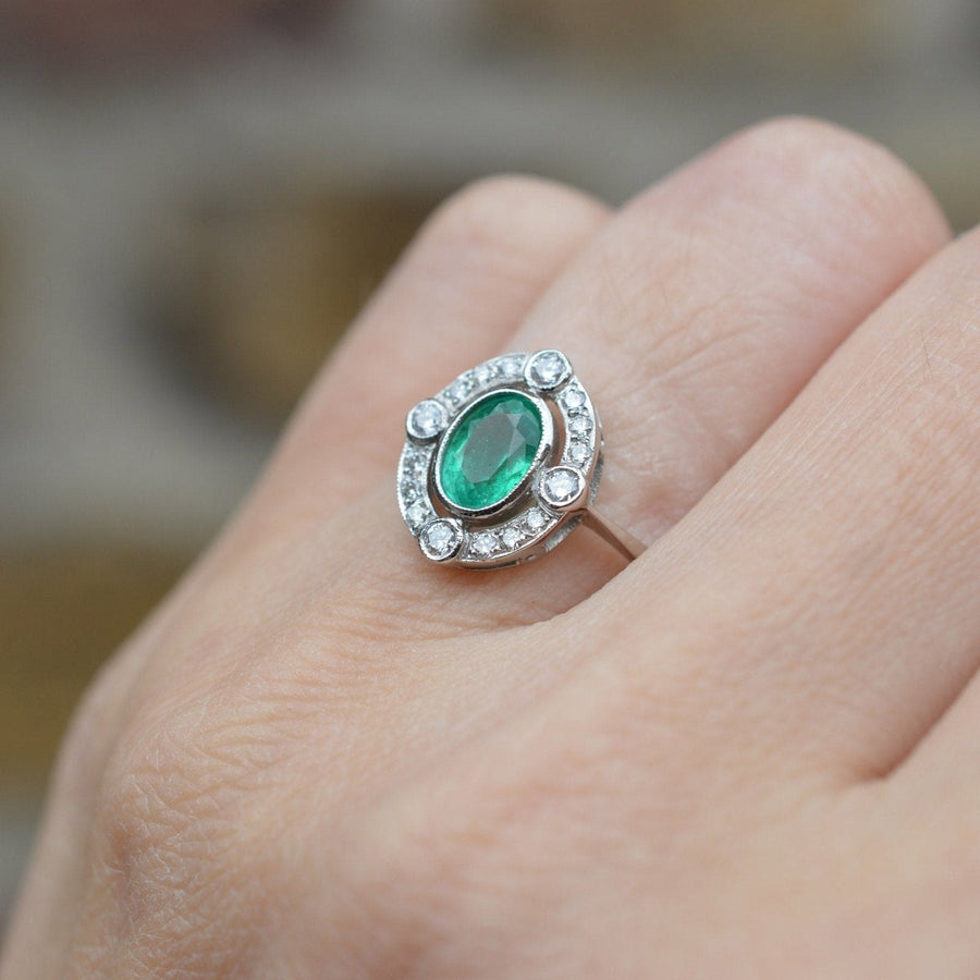 Vintage platinum emerald and diamond cluster ring on a hand, featuring an oval emerald surrounded by small and large diamonds.