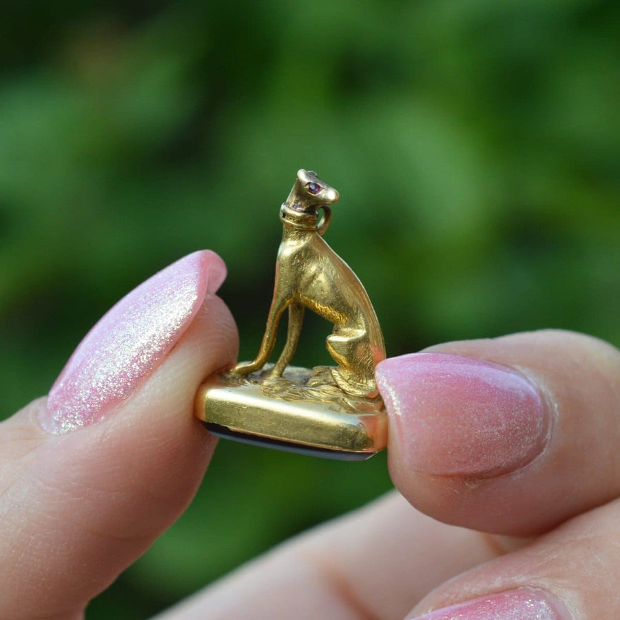 Goldener Whippet-Anhänger mit roten Steinaugen aus dem 19. Jahrhundert, mit einem Sardonyx-Siegelanhänger, in der Hand präsentiert.