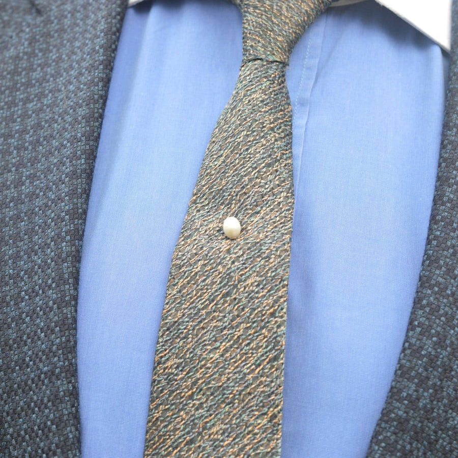 Close-up of a tie pin featuring a natural pearl on a patterned tie and shirt, showcasing vintage elegance.