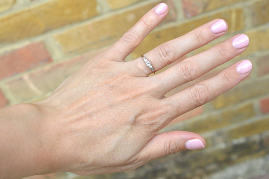 Eleganter Art-Deco-Ring aus 18-karätigem Gold und Platin mit drei Diamanten an einer Hand mit rosafarbenen Nägeln vor einem Hintergrund aus Backstein.