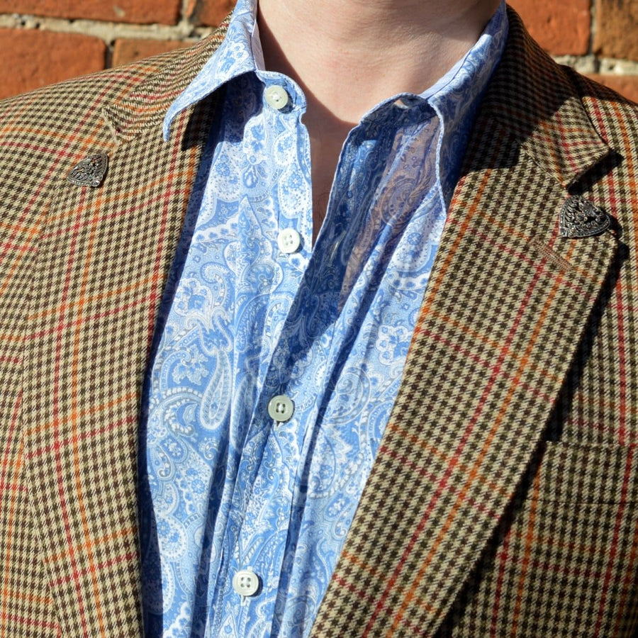 Nahaufnahme eines Mannes, der einen gemusterten braunen Blazer mit Paisley-Hemd und dekorativen Reversnadeln trägt.