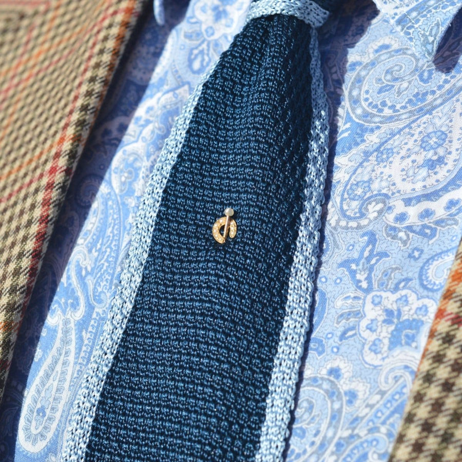 Nahaufnahme einer eleganten blauen Krawatte mit einer goldenen Hufeisenbrosche auf einem gemusterten Hemd und einem Blazer.