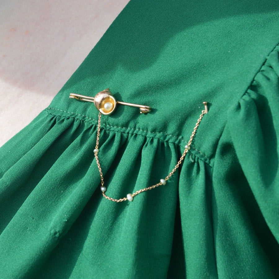 15ct gold bar brooch featuring a seashell and pearl chain, pinned to a green fabric background.