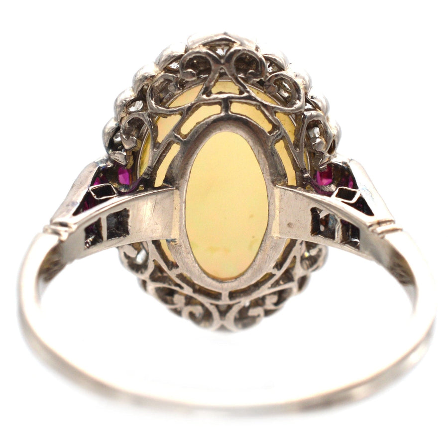 Art Deco 18ct white gold ring featuring an oval yellow opal, surrounded by intricate filigree and accented with Burma rubies.